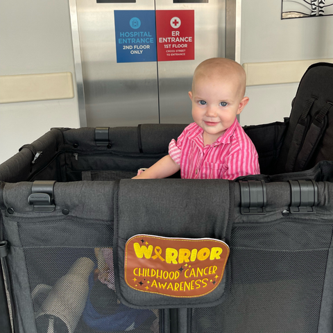 A young child sitting in a Wonderfold wagon outside a hospital entrance, with a 'Warrior: Childhood Cancer Awareness' patch on the front