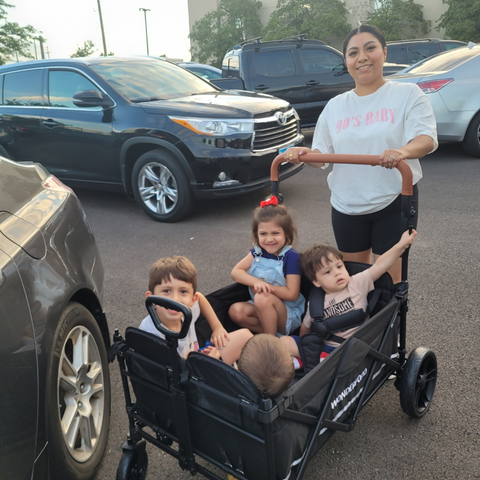 A mother smiling as she pushes four young children in a Wonderfold X4 wagon, preparing for a family outing