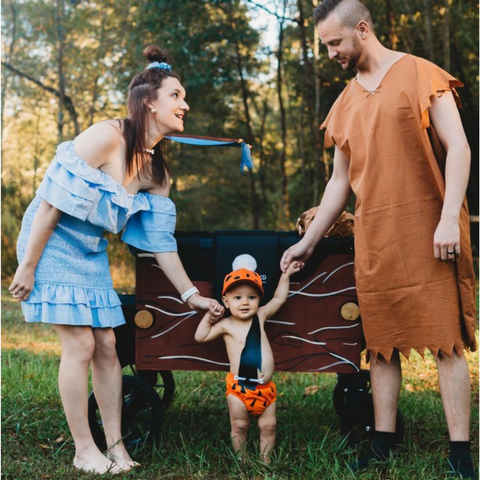 Family dressed as the Flintstones stands in front of their WonderFold wagon transformed into a wooden prehistoric car, featuring a cute baby dressed as Pebbles
