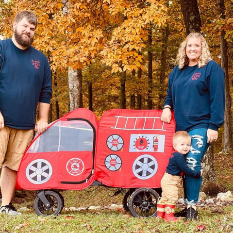 Family dressed as firefighters poses with their WonderFold wagon designed as a red fire truck, surrounded by fall foliage