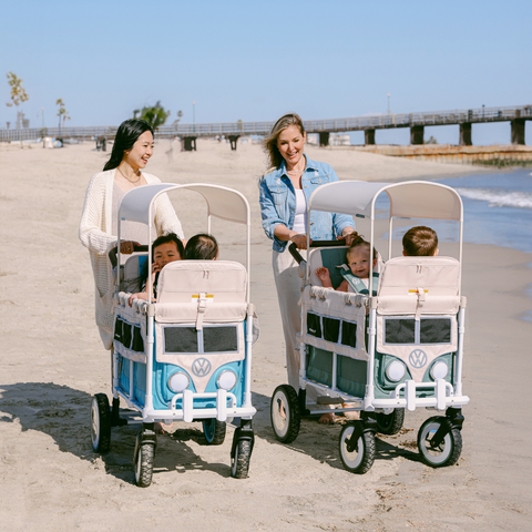 Two women strolling along the sandy beach while pushing Volkswagen-themed WonderFold wagons, each carrying excited children inside