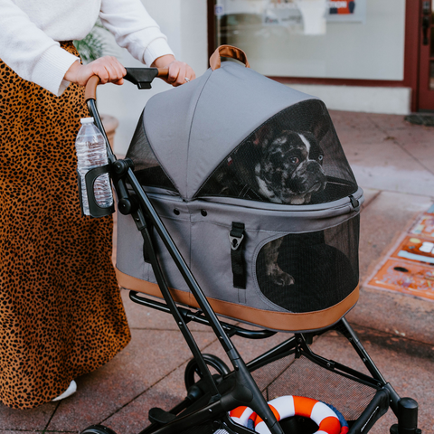 A stylish pet stroller with a mesh canopy carrying a French Bulldog, pushed by a person wearing a leopard-print skirt on a city sidewalk.
