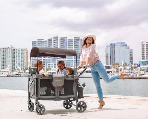 Mom playfully pushes WonderFold stroller wagon with kids by the marina in a cityscape setting