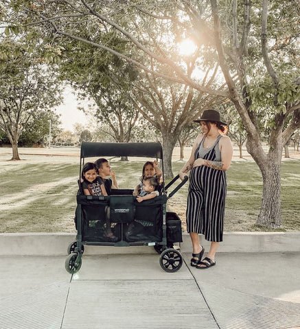 Pregnant mom smiling beside WonderFold stroller wagon carrying three children at a park
