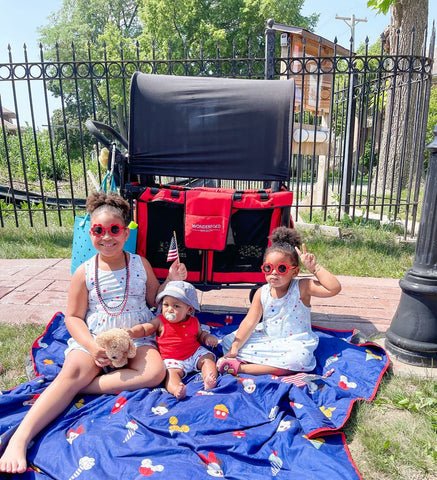Two young girls enjoying a picnic on a patriotic-themed blanket with a red WonderFold stroller wagon in the background.