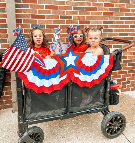 Three children sitting in a WonderFold stroller wagon decorated with American flags and red, white, and blue banners for a patriotic celebration.