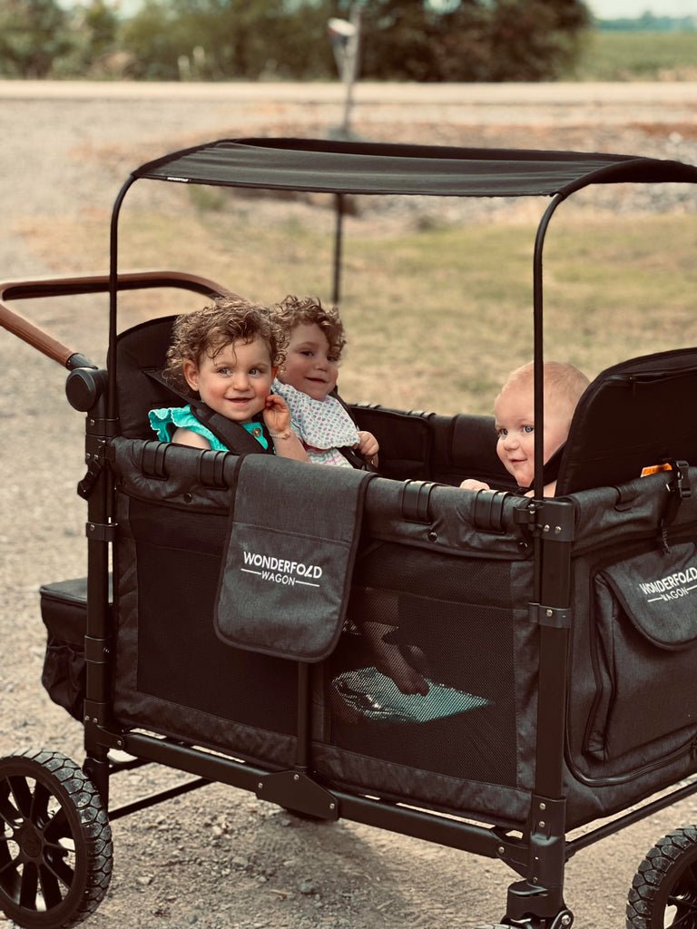 Three smiling toddlers riding in a WonderFold W4 Luxe stroller wagon outdoors
